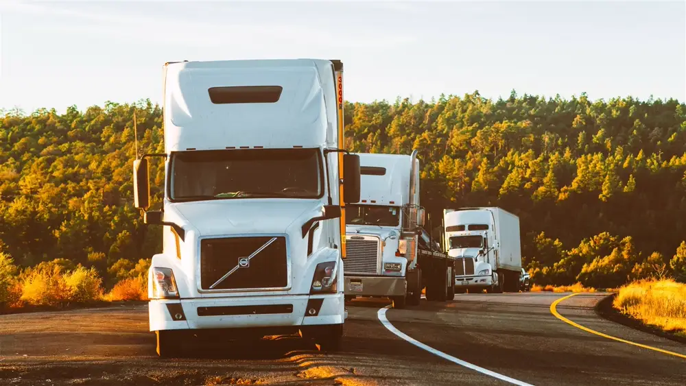 A line of white semi-trucks driving along a highway at sunset